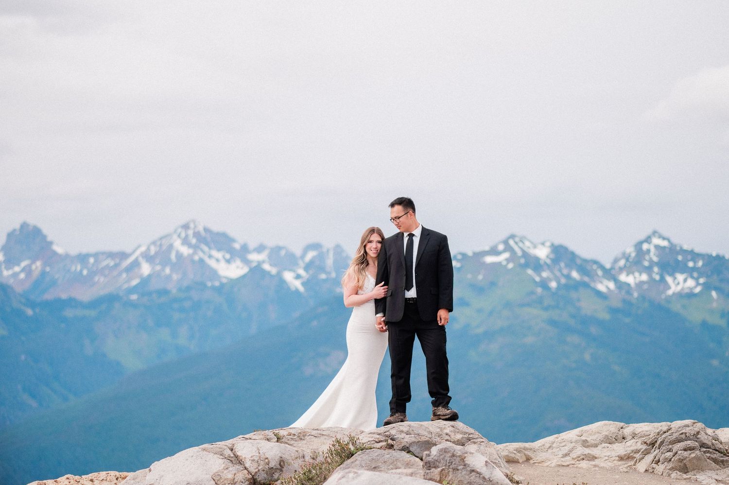 bride and groom snuggling on top of a mountain with blue snow capped mountains behind them. Photo by Washington Elopement Photographer.
