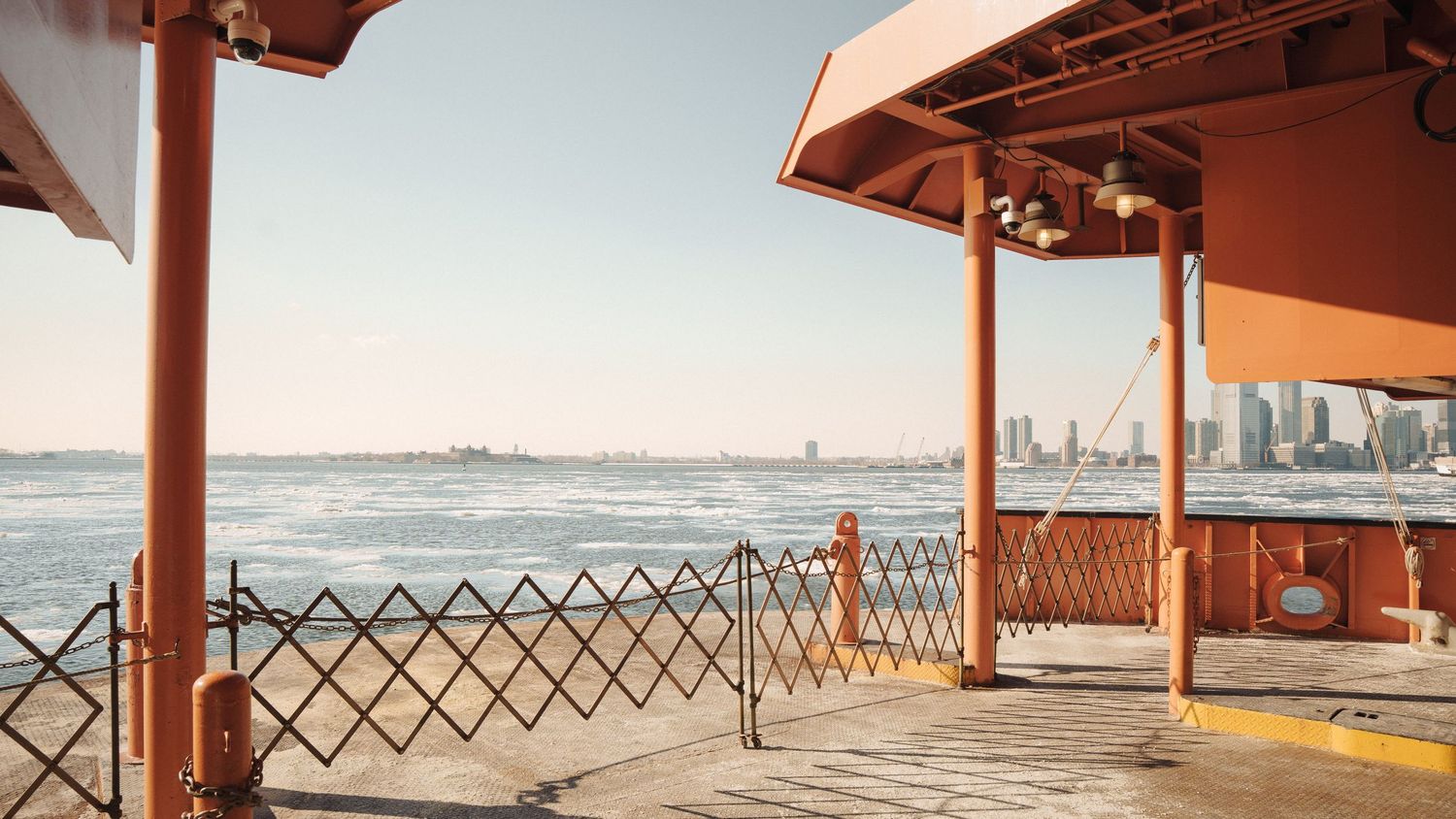 Orange ferry terminal deck overlooking New York Harbor with city skyline in the background on a clear day.