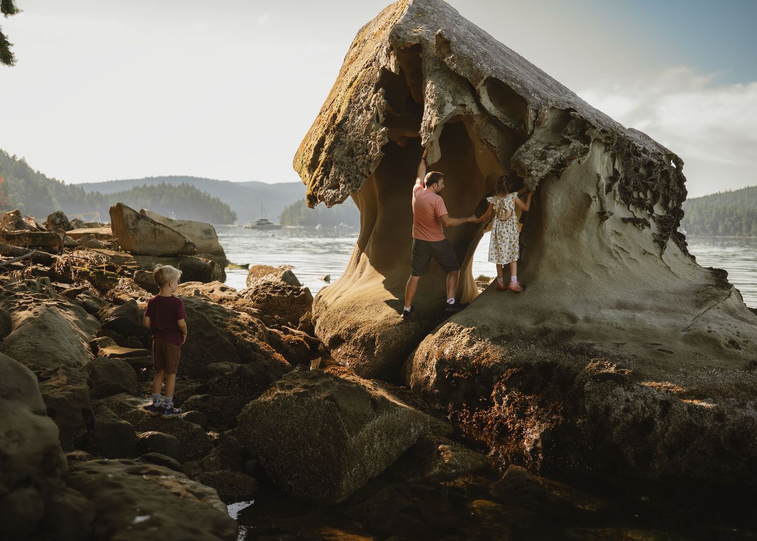 People exploring a large eroded sandstone rock formation on a rocky shoreline surrounded by forested hills.