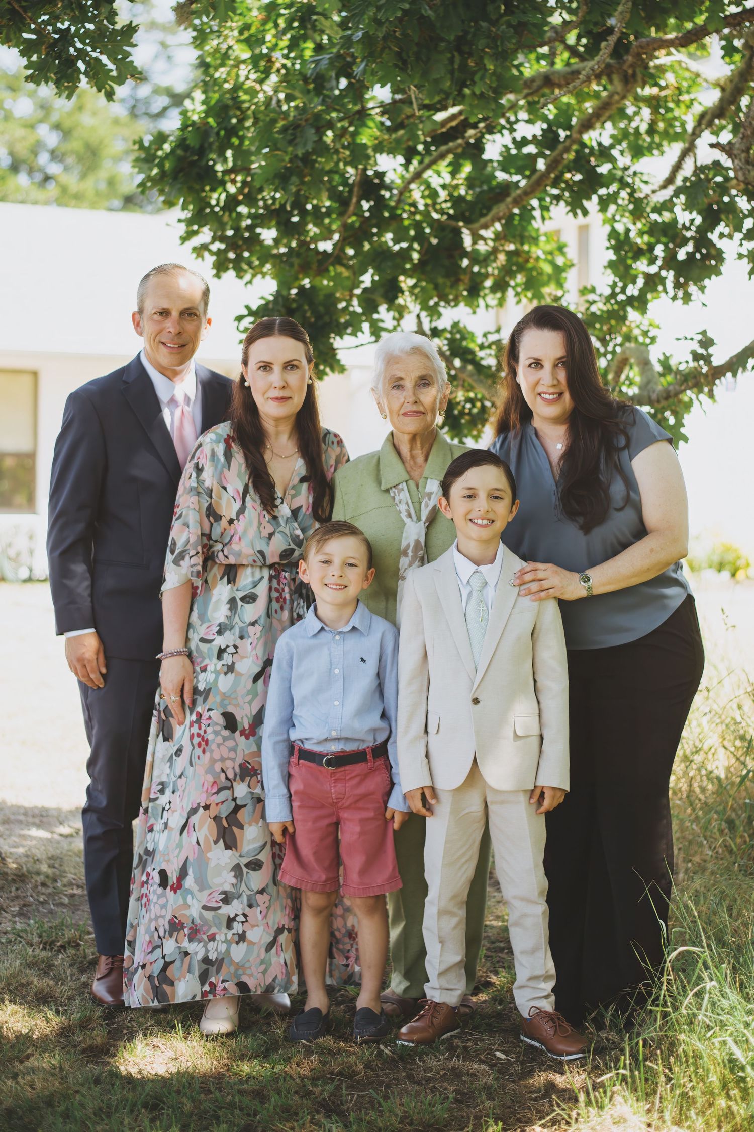 Family group portrait outdoors under green trees on sunny day with floral dress and formal attire.
