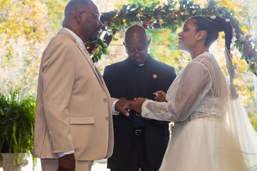 A wedding ceremony takes place outdoors with multiple people gathered in formal attire during a sunny day.