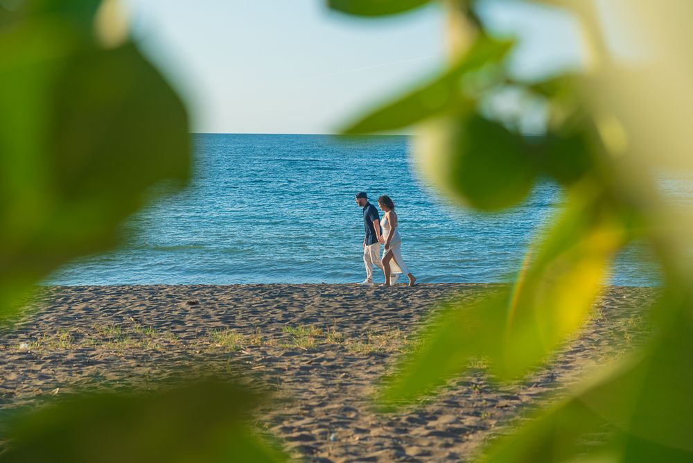 A couple stands together on a rocky beach shore with turquoise ocean waters, framed by green leaves in the foreground.
