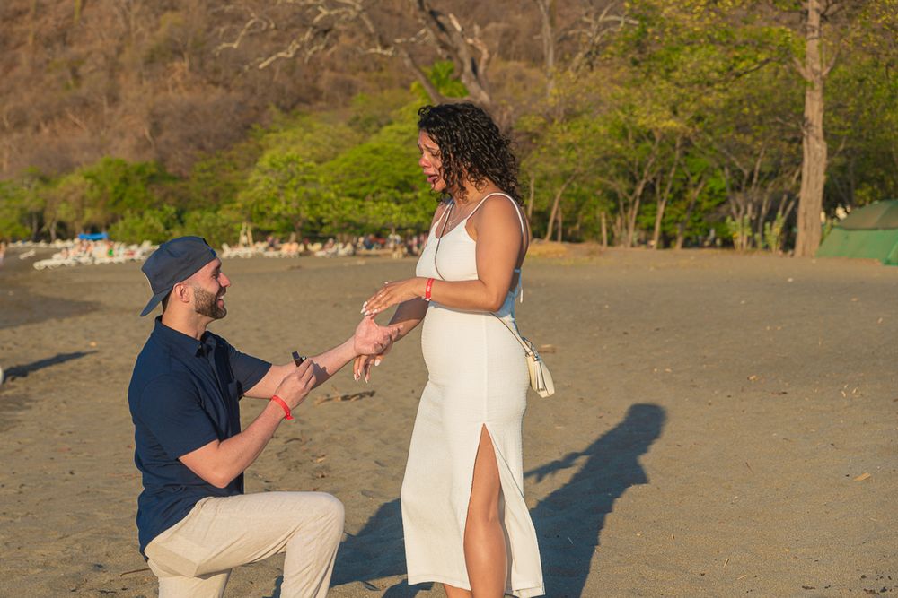 A beach proposal scene unfolds as someone kneels on the sandy shore at sunset.