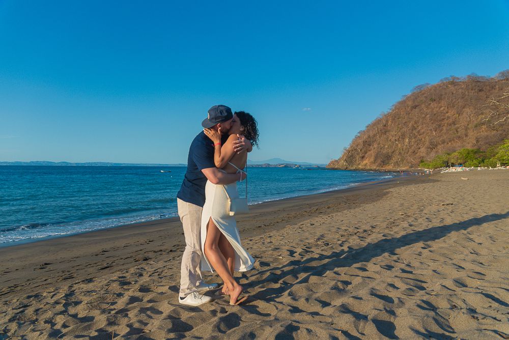 A couple shares a romantic moment on a scenic beach with mountains and blue ocean in the background.