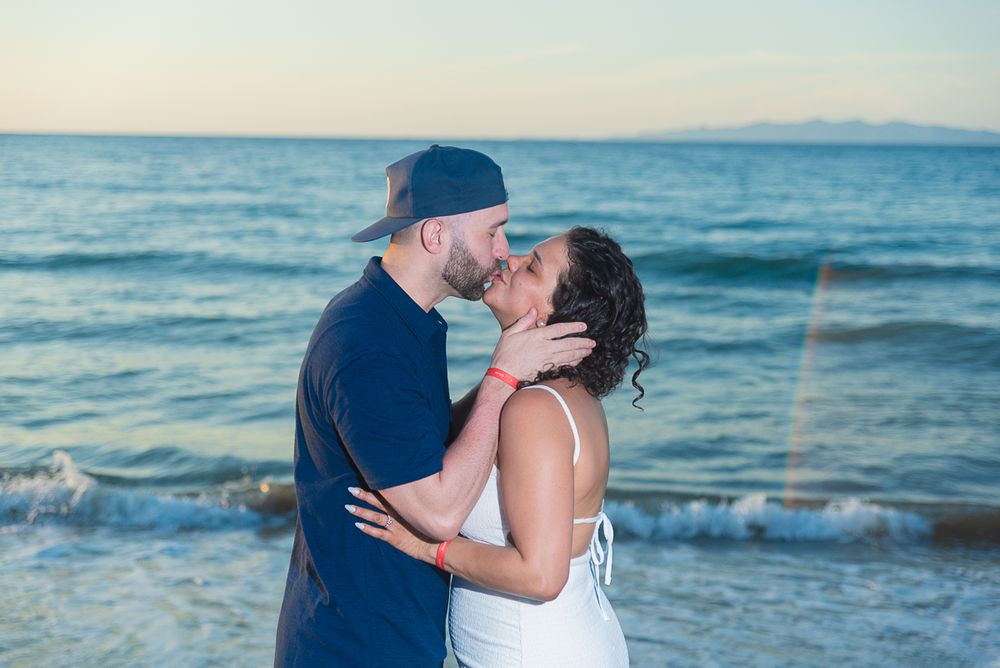 A couple shares a romantic moment on a beach with mountains visible in the background.