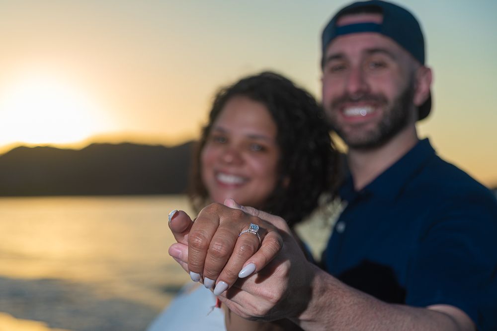 A couple holds up their hands to show off their wedding rings against a beautiful sunset backdrop at the beach.
