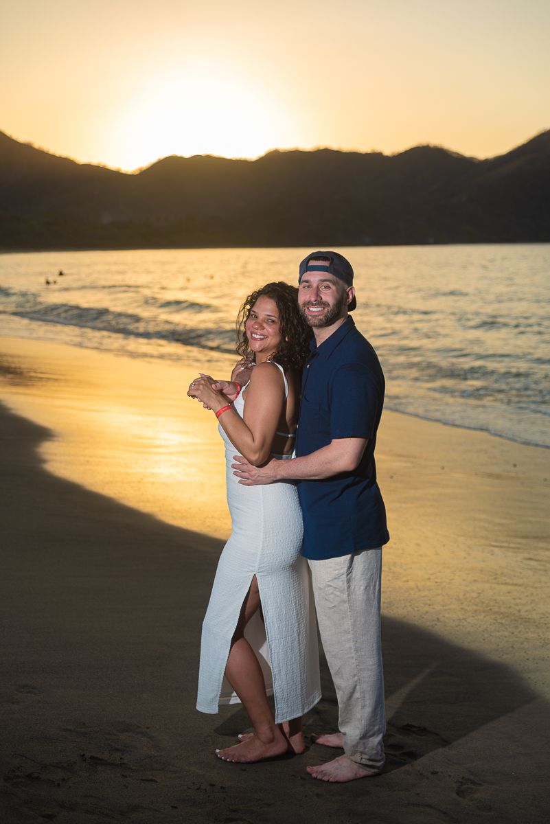 A couple embraces on a beach at sunset with golden light illuminating the shoreline and mountains in the background.