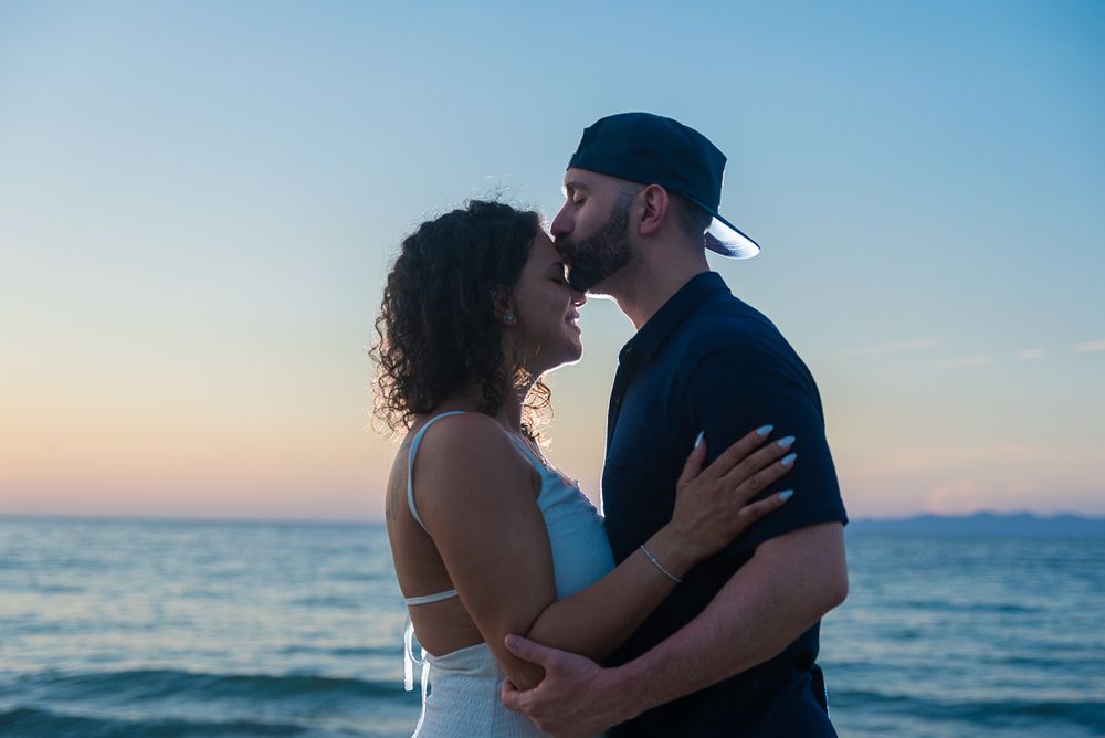 A romantic couple embraces at sunset by the ocean with a soft blue and orange sky in the background.