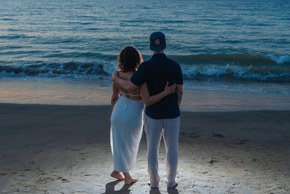 A couple in white outfits stands together watching waves on a beach at dusk.