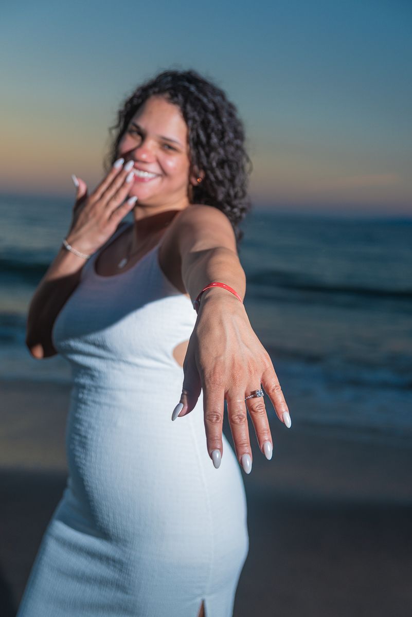 Expectant mother in white dress poses joyfully at sunset on beach during maternity photoshoot.