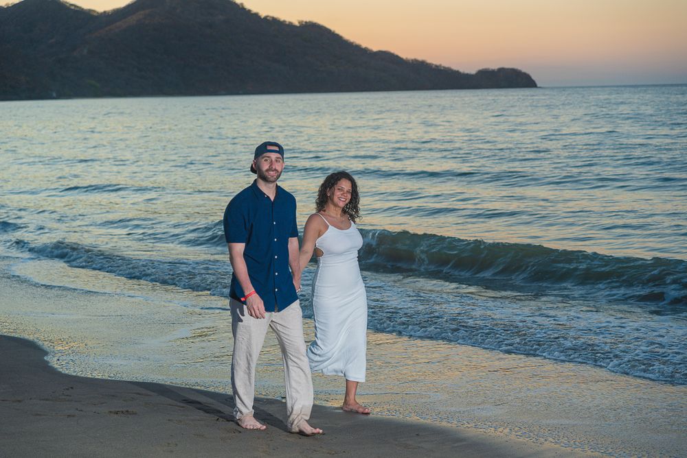 A couple walks hand in hand along a scenic beach at sunset with mountains and ocean waves in the background.