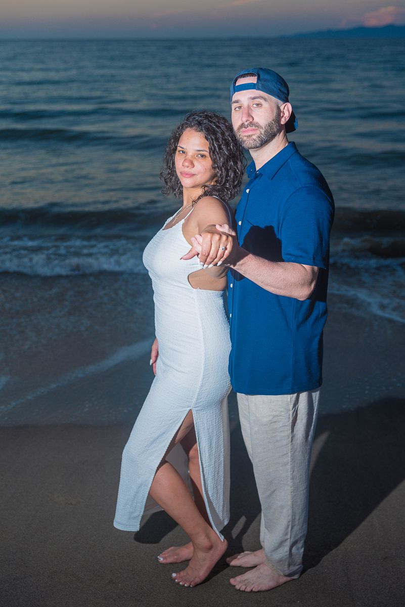 A couple dances barefoot on the beach at dusk, wearing white and blue casual attire by the ocean waves.