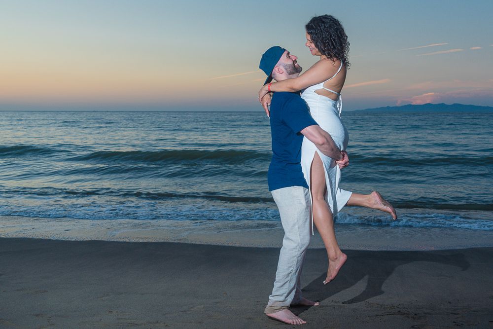 Romantic couple embraces on a sandy beach at sunset with ocean waves in the background.