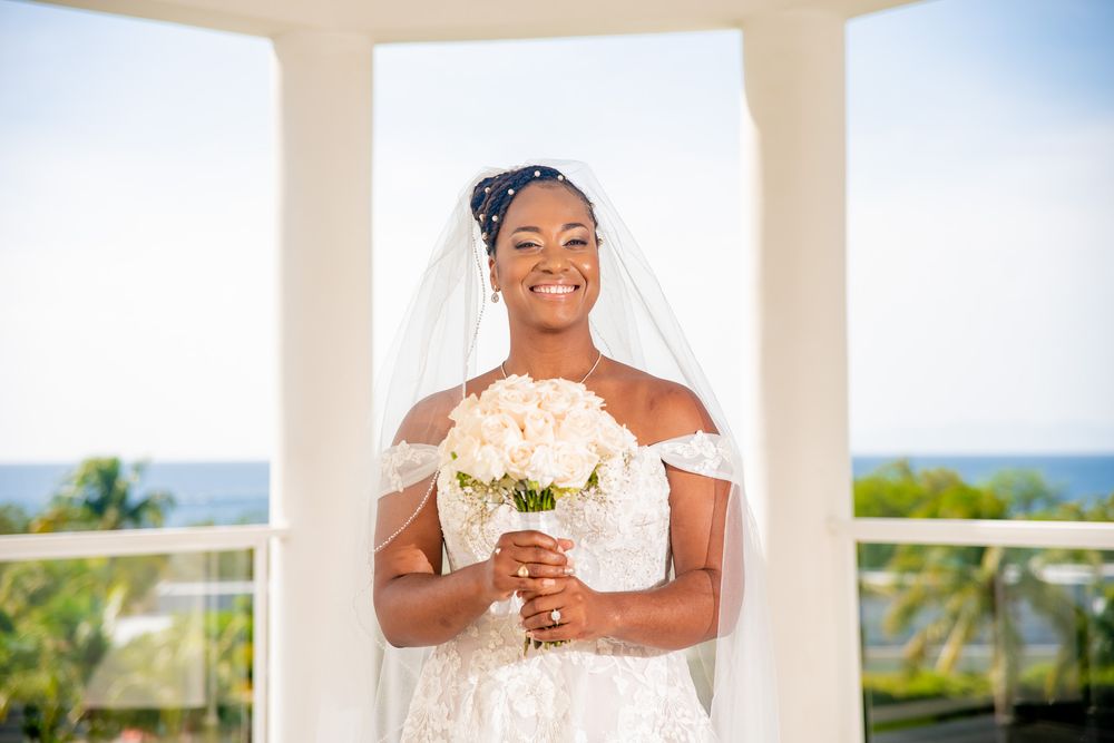 Smiling bride in white wedding dress holds bouquet while standing between white pillars with ocean view in Riu Palace