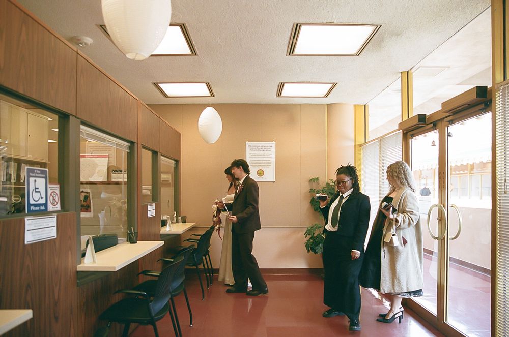 People wait in a vintage medical office waiting room with wood paneling, fluorescent lights and retro furniture from the 1970s.