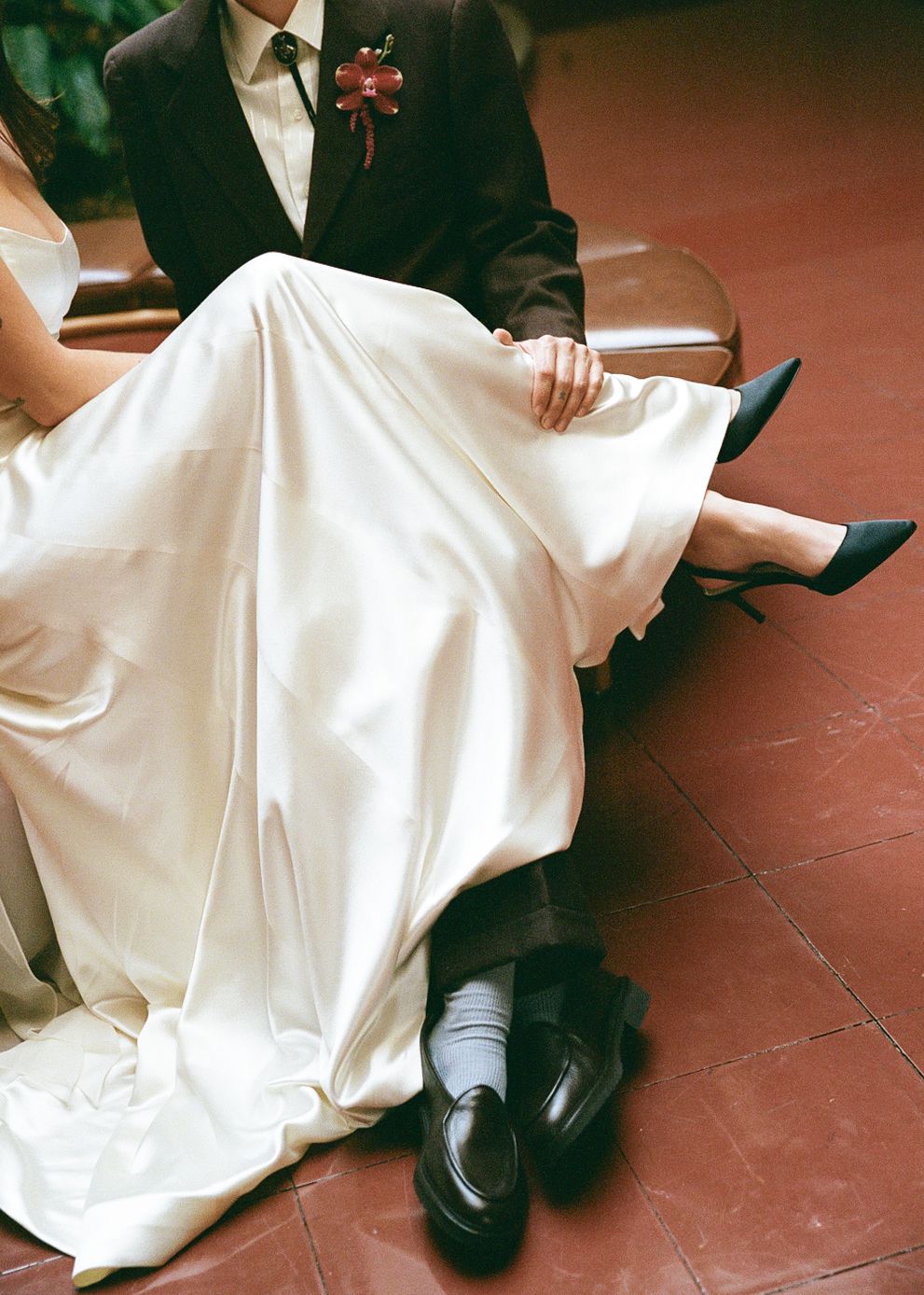 Close up of wedding dress and formal shoes as couple sits together on red floor.