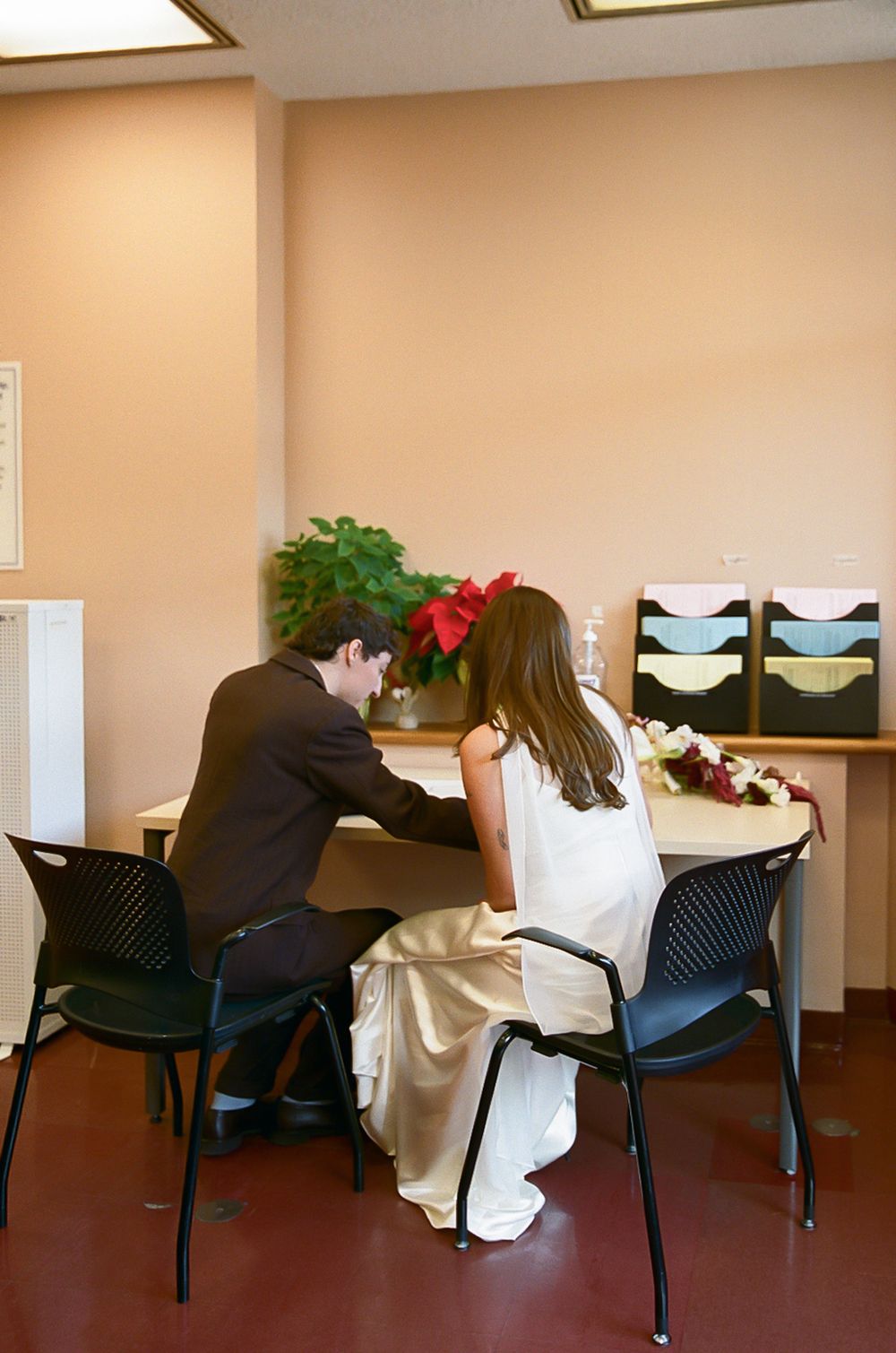 A couple in wedding attire sits together at a table with a poinsettia plant in an office setting.