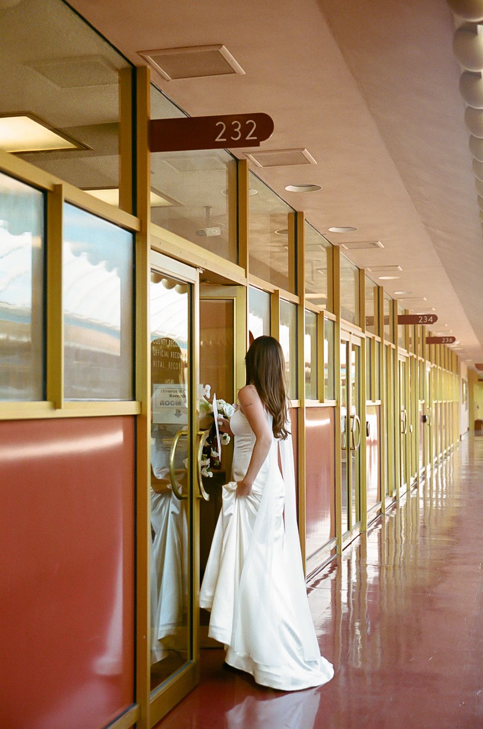 A figure in a white wedding dress stands in a retro-style hallway with red vinyl booths and room number 232.