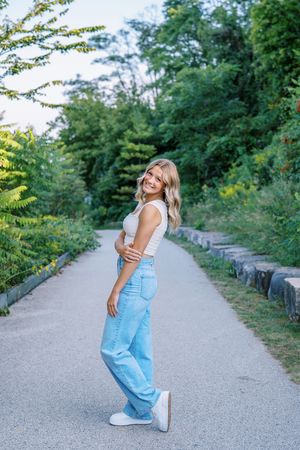 A joyful summer portrait on a paved path surrounded by lush greenery wearing light blue jeans and white top.