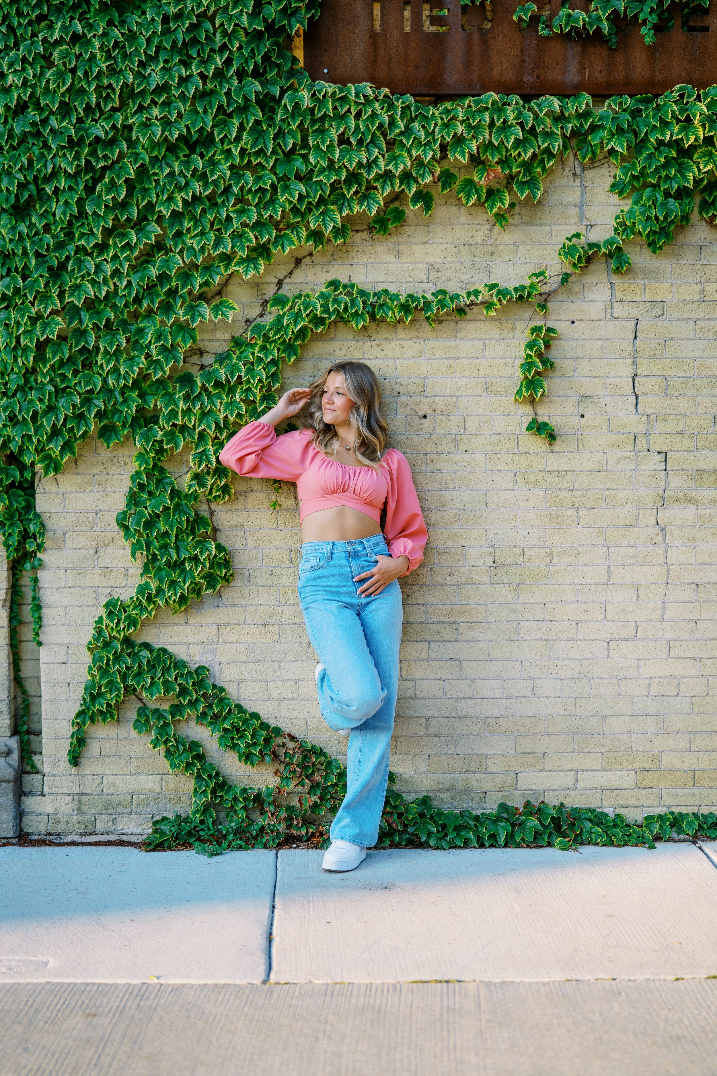 Fashion photoshoot against ivy-covered brick wall showing casual summer style in light denim and pink top.