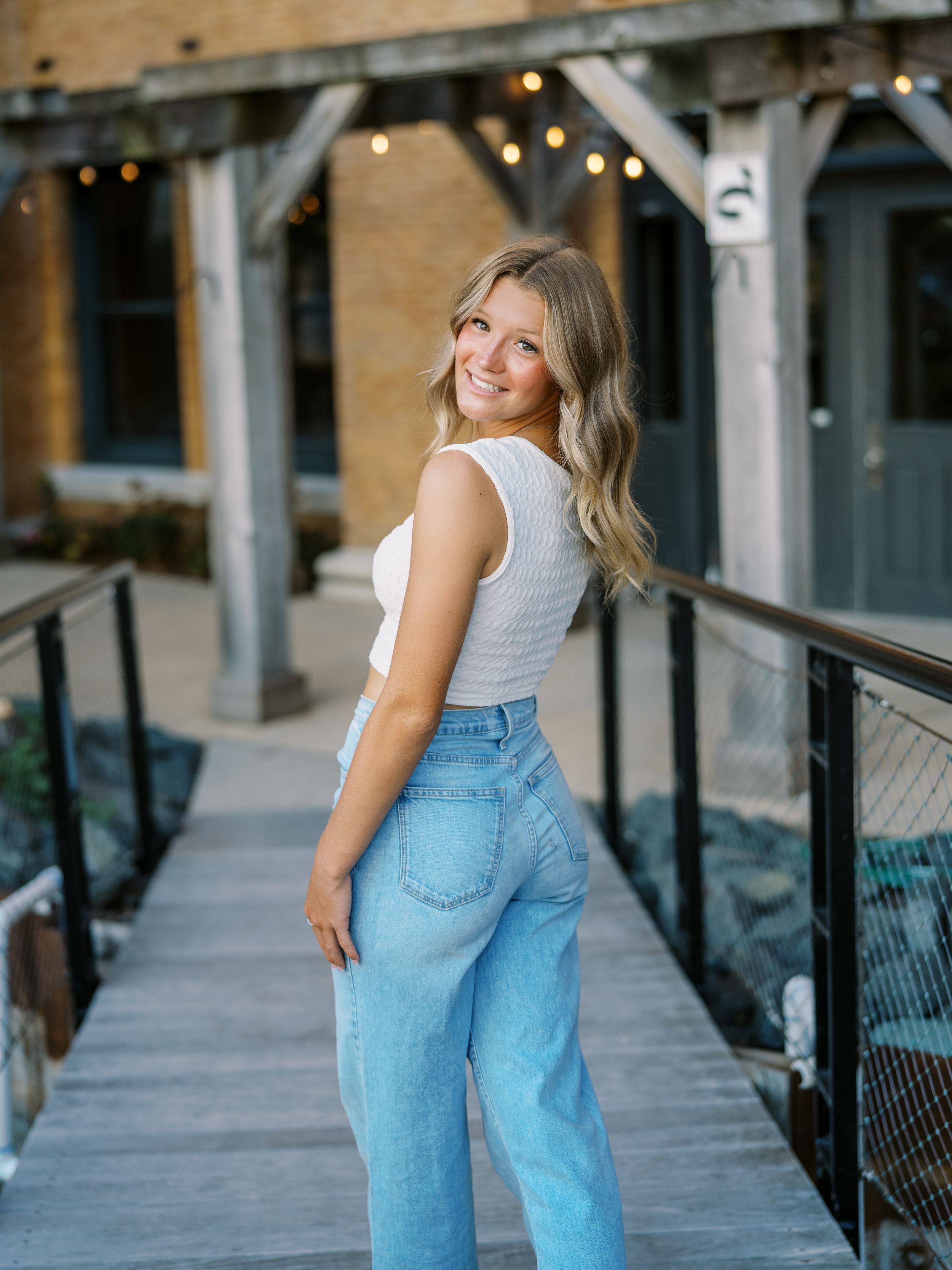 Stylish pose on wooden deck with brick building and string lights in background.
