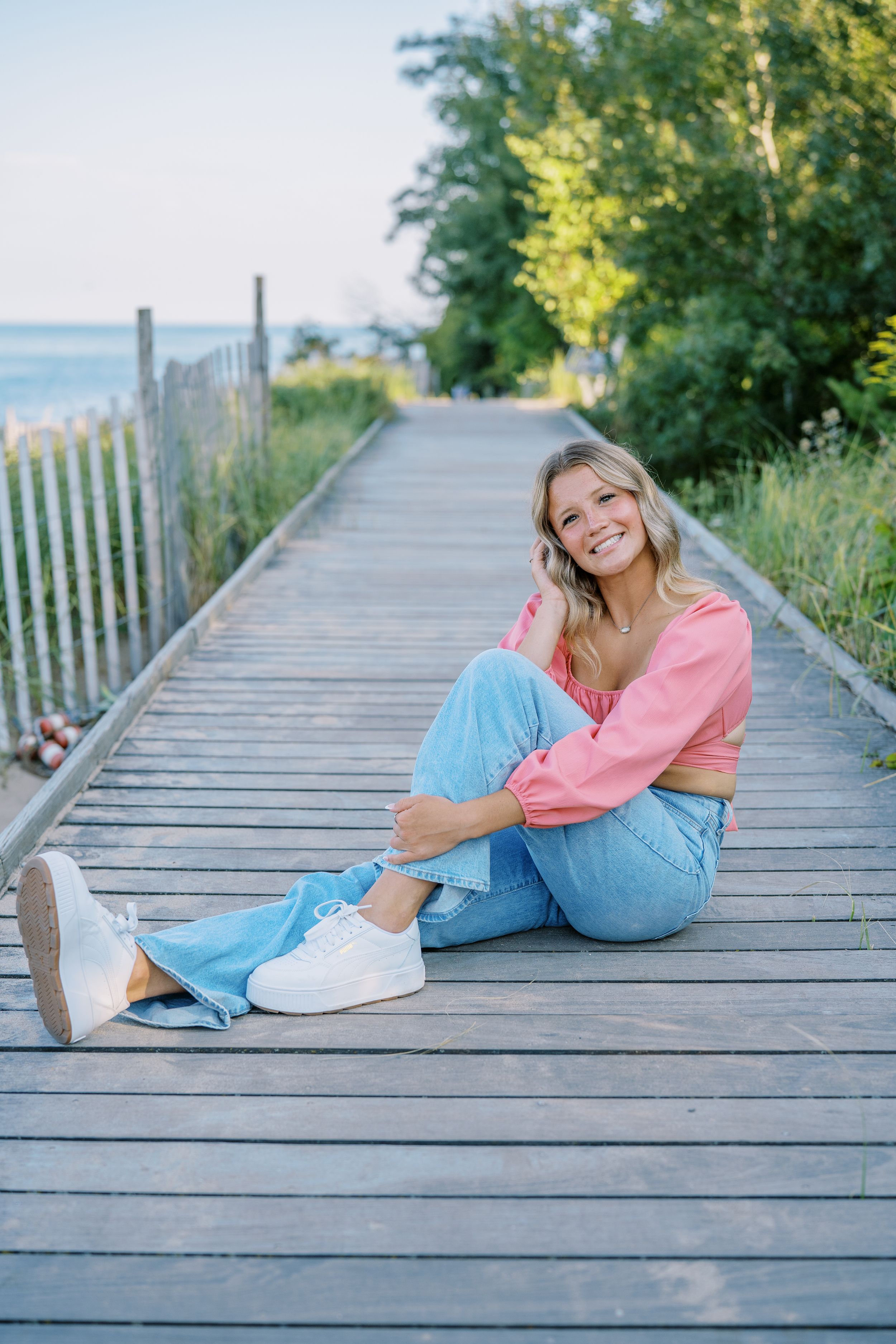 Casual portrait sitting on a wooden boardwalk by the ocean wearing pink sweater and blue jeans.