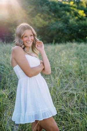 Candid outdoor portrait in a white summer dress standing in tall grass during golden hour.