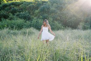 Ethereal portrait in a flowing white dress standing among tall grasses at sunset.