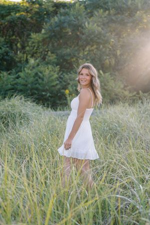 Summer portrait in a white sundress standing in a field of tall grass with sunlight behind.