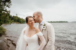 A couple shares an intimate moment on a rocky beach shoreline with a lake and trees in the background.