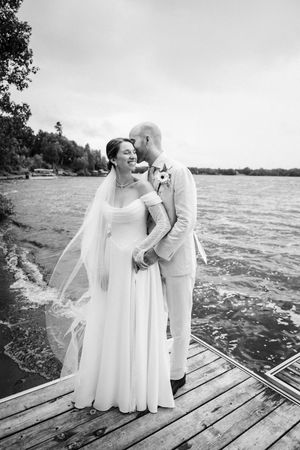 A romantic black and white wedding photo of a couple embracing on a wooden dock by a lake.
