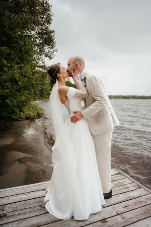 A romantic wedding couple embraces on a wooden dock by a lake shore on an overcast day.
