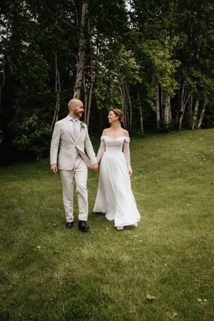 A couple in white wedding attire walks together on a grassy hillside surrounded by trees on a sunny day.