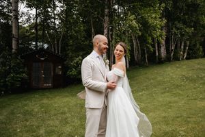 A couple shares a romantic moment on a grassy hillside with birch trees and a rustic cabin in the background.
