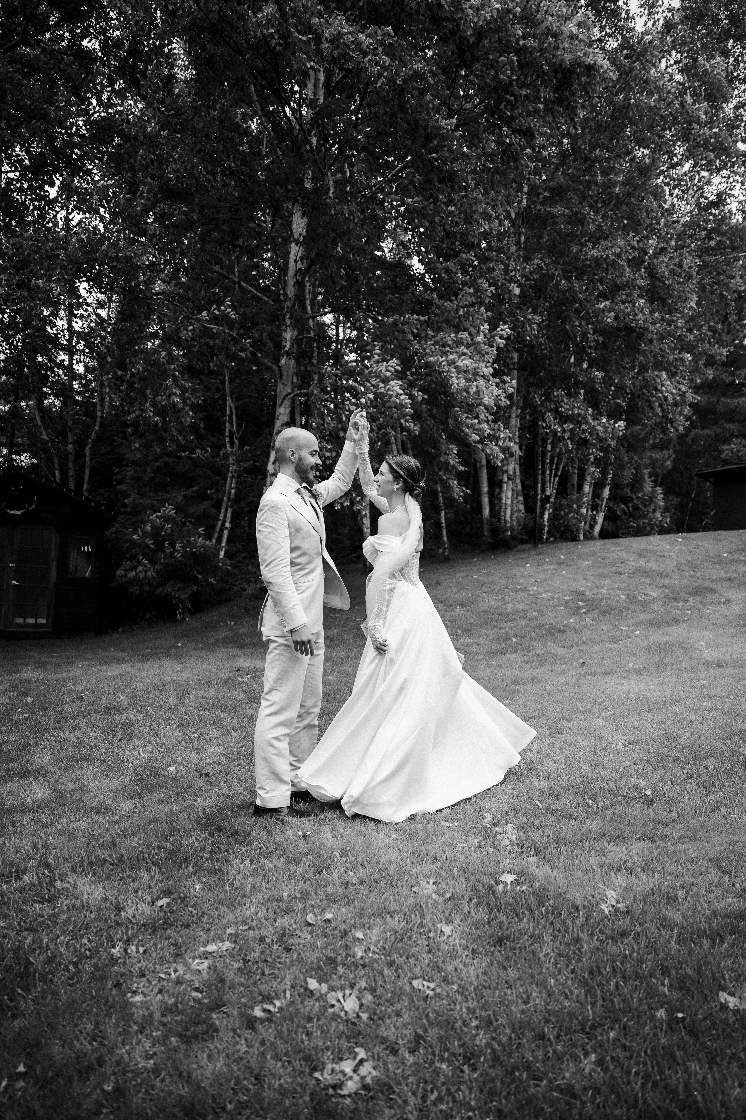 A couple shares a romantic dance moment on a grassy lawn surrounded by trees in black and white photography.