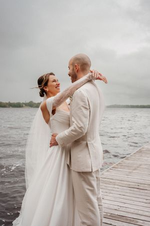A couple shares an intimate moment on a wooden dock by a lake on their wedding day in light colored attire.
