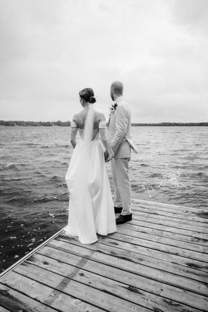 A couple stands on a wooden dock overlooking a lake during their black and white wedding photoshoot.