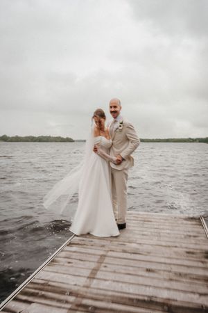 A couple in wedding attire stands on a wooden dock over choppy waters on an overcast day.