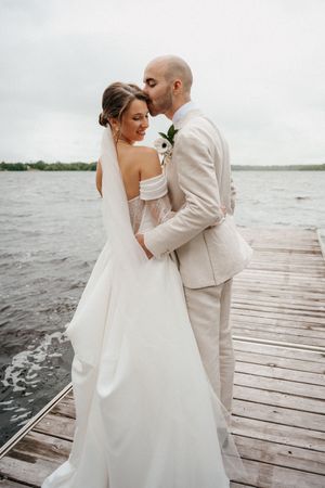 A romantic wedding couple embraces on a wooden dock by the water, with the bride in white and groom in beige.