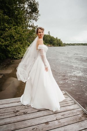 A bride in a flowing white gown with long sleeves poses gracefully on a wooden dock by the water.