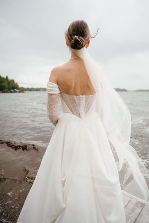 Flowing white wedding dress with lace details and sheer back photographed against a moody waterfront backdrop.