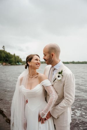 A romantic wedding couple embraces by a lakeside on an overcast day, wearing elegant cream and white attire.