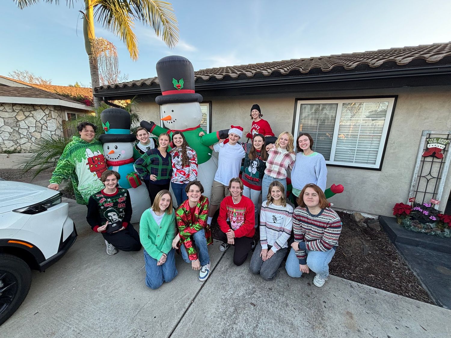 A group of teenagers in holiday sweaters pose outdoors with an inflatable snowman decoration.