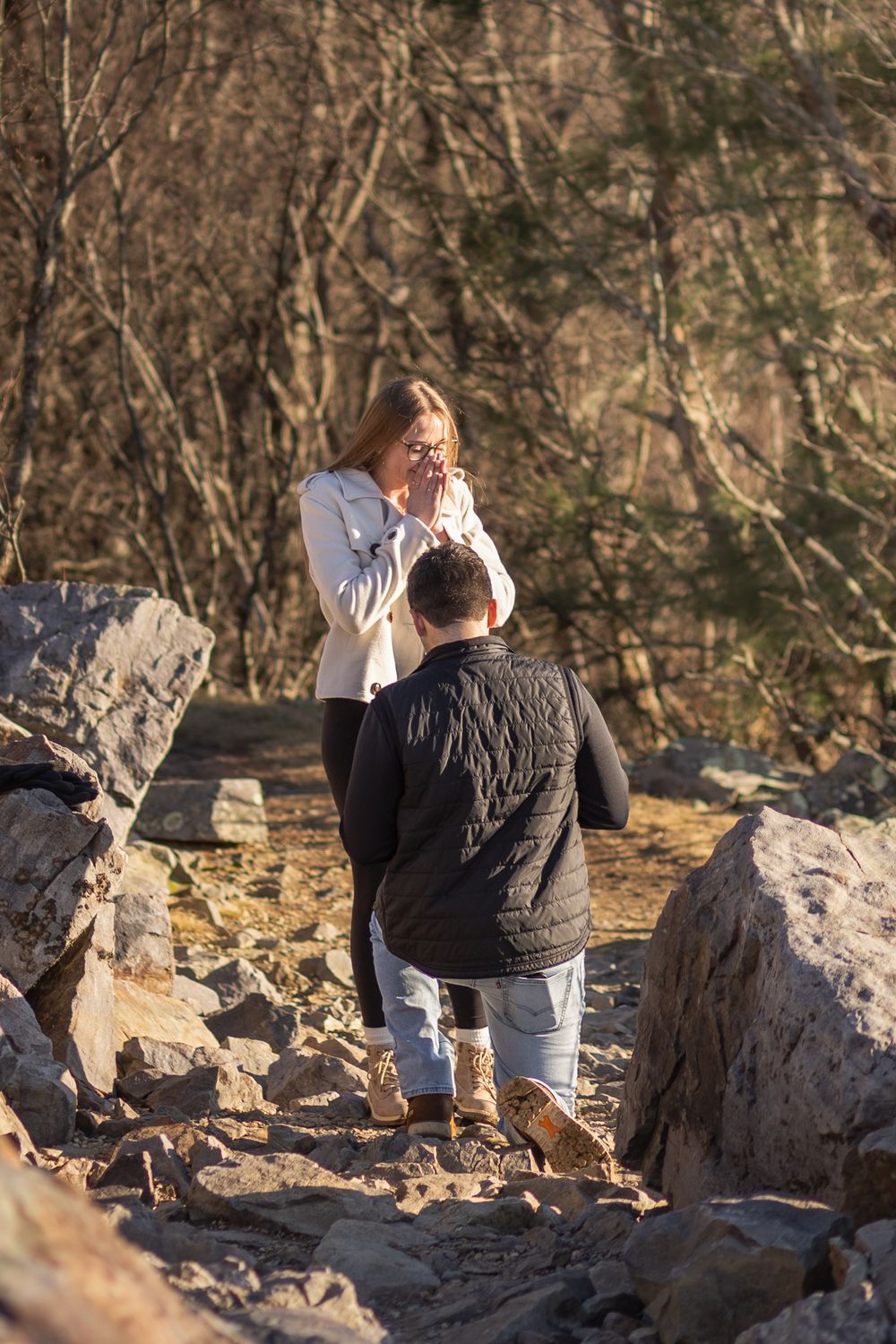 A couple shares a special moment on rocky terrain during what appears to be a surprise proposal outdoors.