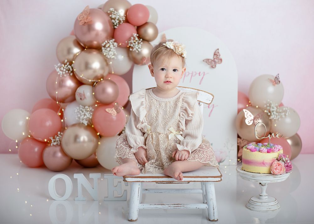 First birthday celebration setup with rose gold and pearl balloon arch, white bench, and decorative cake on pedestal.