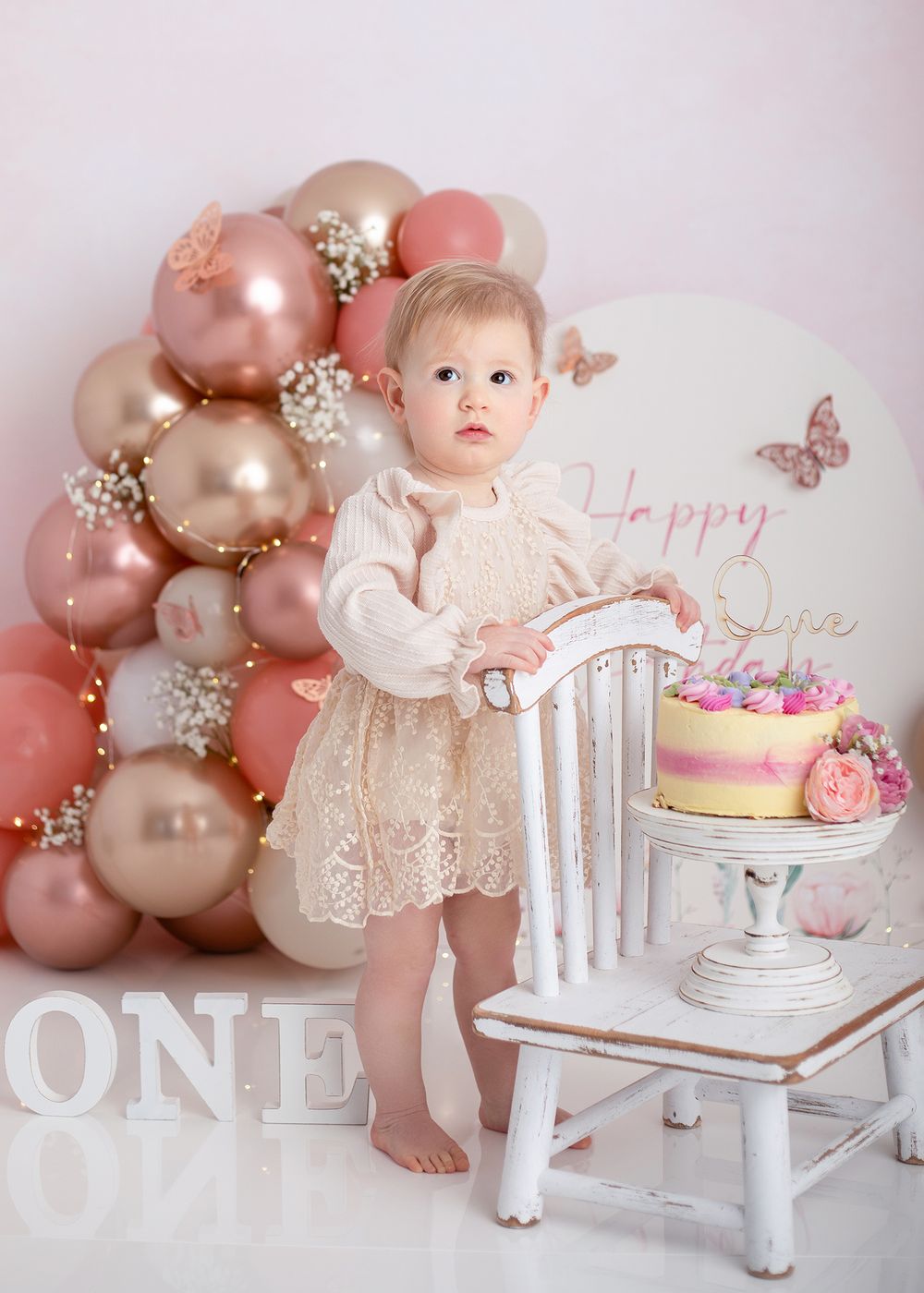 Pink and gold balloon arch decorates a white chair and cake setup for a first birthday celebration photoshoot.