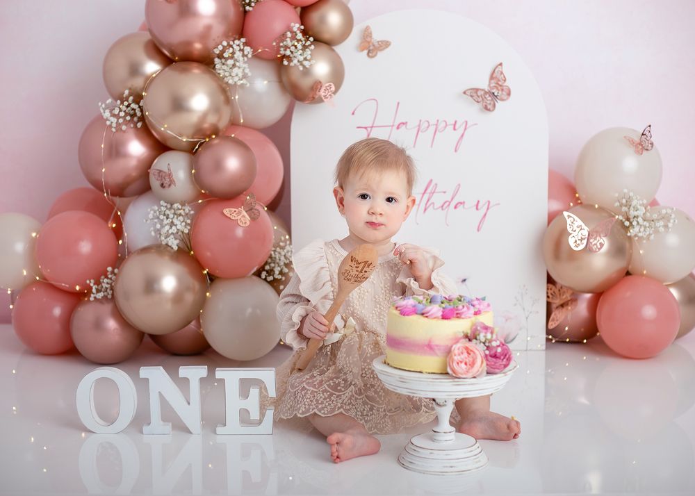 Pink and gold balloon garland decorates a first birthday party setup with a white cake and delicate butterfly accents.
