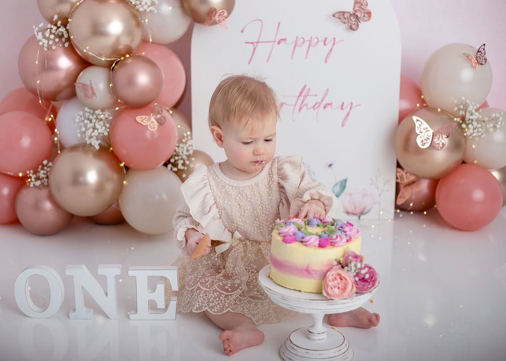 First birthday celebration setup with rose gold balloon arch, white 'ONE' letters, and a colorful tiered cake on a pedestal.