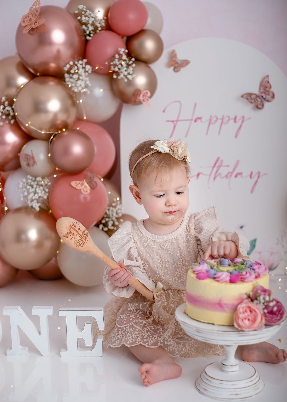 A festive first birthday setup with pink and gold balloon arch, decorative butterflies, and a pink floral cake on white pedestal.