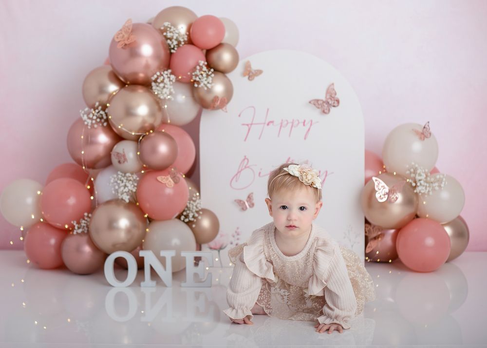Elegant first birthday photo session with rose gold and pink balloon decorations against white backdrop.